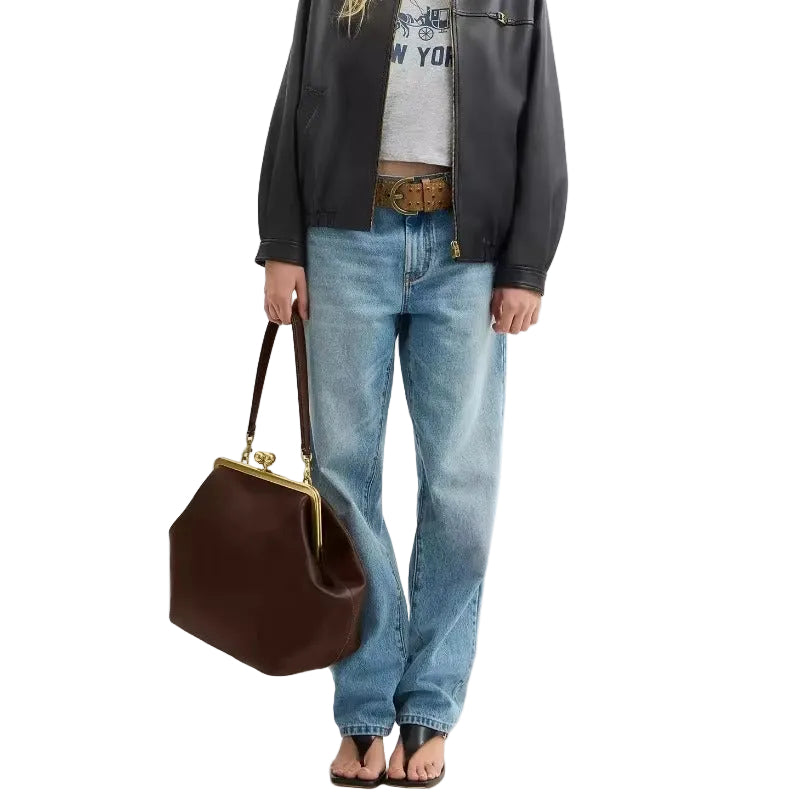 A model wearing a black leather jacket, light-wash blue jeans, and a gray graphic t-shirt, holding a dark brown leather handbag with a gold-tone frame against a plain white background.