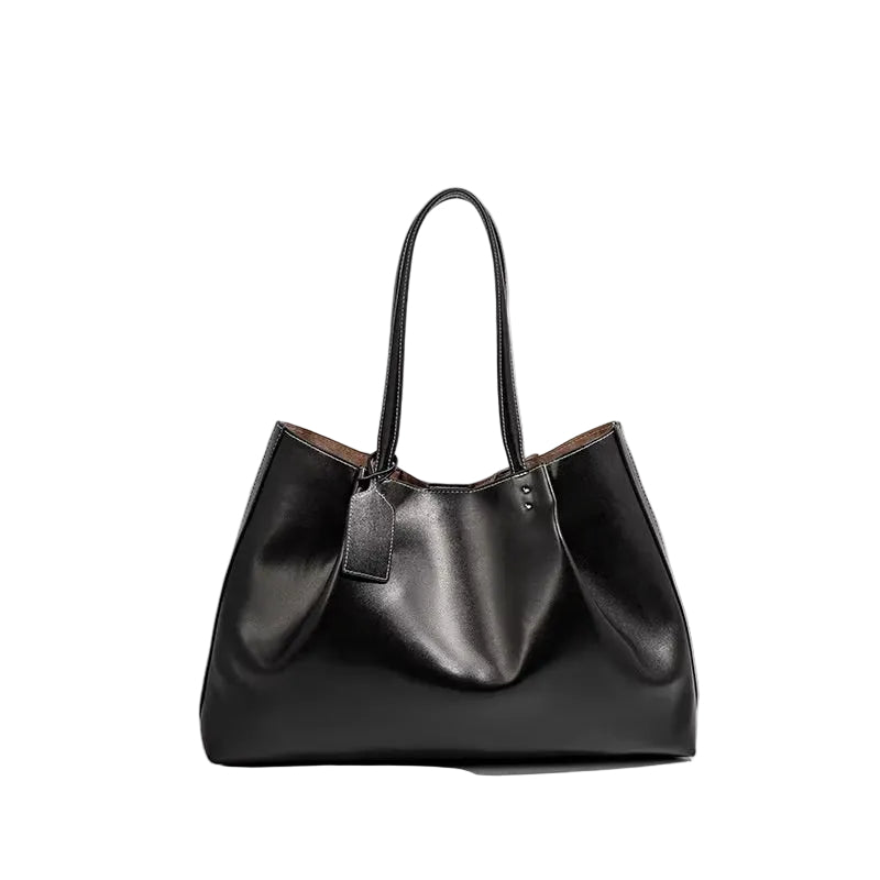 A black leather slouchy tote bag with dual shoulder straps and a hanging leather tag, photographed on a light gray background with a soft shadow.