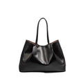 A black leather slouchy tote bag with dual shoulder straps and a hanging leather tag, photographed on a light gray background with a soft shadow.